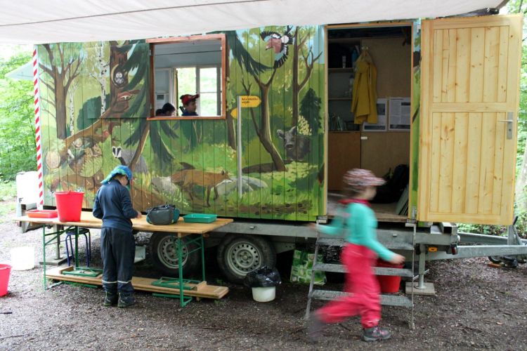 Der Naturdetektiv-Wagen steht im Wald, davor aufgestellt sind verschiedene Forschungsutensilien wie Kescher und Kübel.